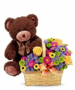 A wicker basket full of bright cheery flowers with a cute teddy bear.